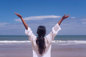 Photo of a woman standing on a sunny beach holding her hands up in the air. This photo represents how you can defeat the depression blues with depression therapy in California. 