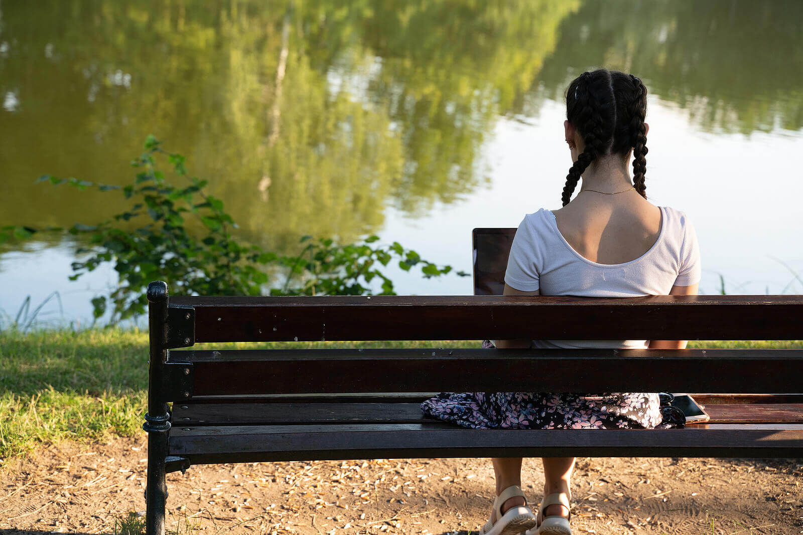 Photo of an Asian American woman sitting on a bench using her laptop by the pond. This photo represents how therapy for brainspotting in California can help you find peace by coping with your trauma.