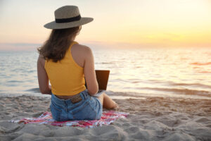 Photo of a woman sitting on a beach during sunset using a laptop. By meeting with an online trauma therapist you can begin to heal from your trauma symptoms. Learn what questions you should ask here.